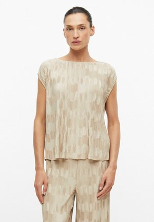 Femme debout face à l'avant, portant un haut beige ample à manches courtes et un pantalon assorti avec un subtil motif géométrique texturé, sur un fond uni.