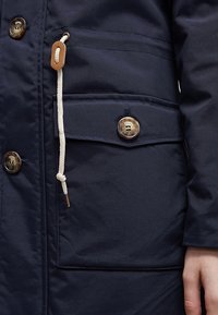 Marinblå jacka med en slät textur, stora knappar, dragsnöre med en träflip, och en framficka med lock.