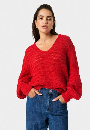 Rød strikket sweater med V-hals, ribbet tekstur og lange ærmer, parret med højtaljede blå denimjeans.