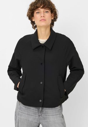 Frau trägt eine schwarze, geknöpfte Jacke mit Kragen über einem gestreiften Hemd und hellgrauen Jeans, stehend mit den Händen in den Taschen.