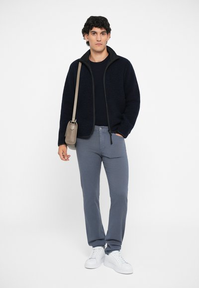 Marineblaue Fleecejacke mit schwarzem Besatz, graue Hose und weiße Sneakers. Eine kleine beige Umhängetasche wird über der Schulter getragen.