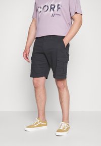Pantalones cortos de carga negros con bolsillos laterales, hechos de una tela duradera. Combinados con una camiseta gráfica de color púrpura claro y zapatillas beige.