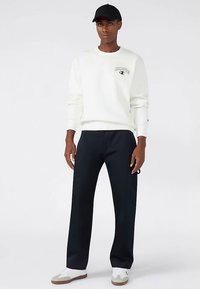 Weißes Sweatshirt mit Logo, schwarze Hose und weiße Turnschuhe. Die Person trägt eine schwarze Kappe und steht vor einem neutralen Hintergrund.