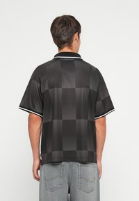 Άνδρας που φοράει μαύρο καρό πόλο μπλουζάκι με λευκή περιθώριο και ανοιχτό μπλε τζιν, εμφανιζόμενος από πίσω σε απλό φόντο.