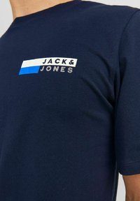 Tumši zils kokvilnas T-krekls ar baltu un zilu taisnstūra logotipu, uz kura redzams teksts "JACK & JONES", īsām piedurknēm un apaļu kakla izgriezumu.