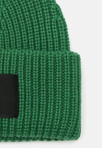 Berretto lavorato a maglia verde con texture a coste e orlo ripiegato. Presenta un'etichetta nera solida sul davanti. Materiale morbido.