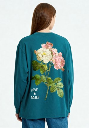 Vrouw met de rug naar de kijker die een blauwgroen shirt met lange mouwen draagt met een roze en witte roos en de tekst "LOVE & ROSES" op de achterkant.