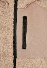 Fleecejackenabschnitt mit weichem, beigem Material, strukturiertem Außenbereich und einer kontrastierenden schwarzen Reißverschlusstasche auf einem glatten, rosa Stoffhintergrund.