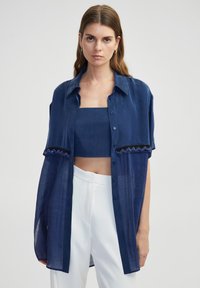 Camisa azul marino de botones y manga corta con textura semitranslúcida, con adornos de cuentas a lo largo del dobladillo, llevada sobre un top corto a juego.