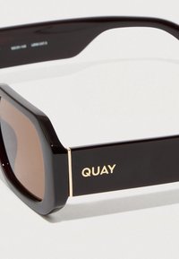 Črna sončna očala s kvadratnim okvirjem, rjavimi stekli in zlatimi poudarki z imenom blagovne znamke "QUAY" na strani. Gladek plastičen material.