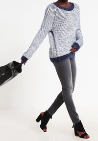 Ljusblå stickad tröja med luddig marinblå kant, grå åtsittande jeans och svarta mocka högklackade stövlar, håller en svart tote-väska.