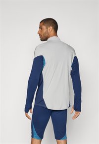 Sportoutfit med en lys grå langærmet bluse med blå og teal detaljer, parret med marineblå shorts. Glat, åndbar stof.