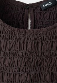 Tissu froncé brun foncé en gros plan avec des coutures horizontales et une seule fermeture par bouton près de l'encolure, montrant l'étiquette de la marque MNG.