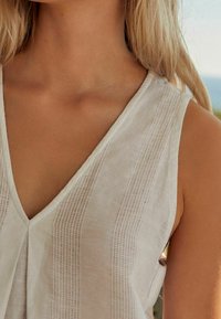 Light beige sleeveless blouse with a V-neck, featuring vertical stripes and textured detailing on the fabric. Simple, elegant design.
