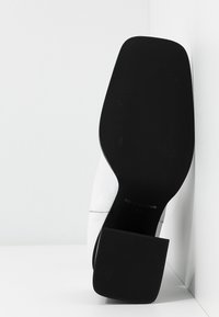 Botte blanche à semelle noire avec talon carré, semelle tournée vers la caméra, positionnée verticalement contre un angle et une surface blancs.