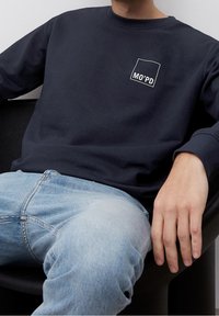 Homme assis portant un sweat-shirt bleu marine avec le logo "MO'PD" et un jean bleu clair, reposant la main droite sur l'accoudoir noir d'une chaise.