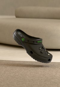 Zwarte klompen met groene accenten, voorzien van knoopachtige details op het bovenwerk. Gemaakt van lichtgewicht materiaal, met een gestructureerde zool en een ademend ontwerp.