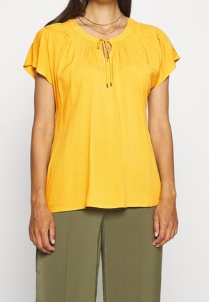 Woman wearing a short-sleeve yellow blouse with a tie neckline and olive green pants against a plain background.