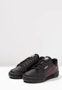 Czarne buty sportowe z gładkiej skóry, z kontrastującą czerwoną paskiem, czarnymi sznurowadłami i białym logo na języku.