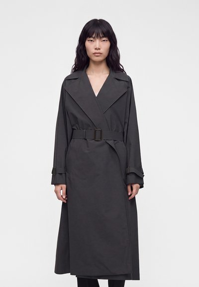 Femme aux longs cheveux noirs portant un trench-coat gris foncé ceinturé, debout devant un fond blanc uni.
