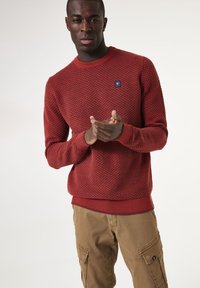 Roter strukturierter Strickpullover mit rundem Ausschnitt und gerippten Bündchen. Verfügt über ein kleines blaues Logo-Patch. Kombiniert mit hellbraunen Cargo-Hosen.