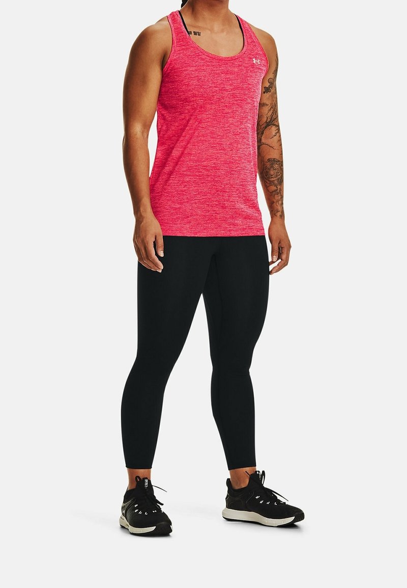 Personne portant un débardeur de sport rose sans manches, un legging noir et des chaussures de course noires, debout devant un fond clair uni.