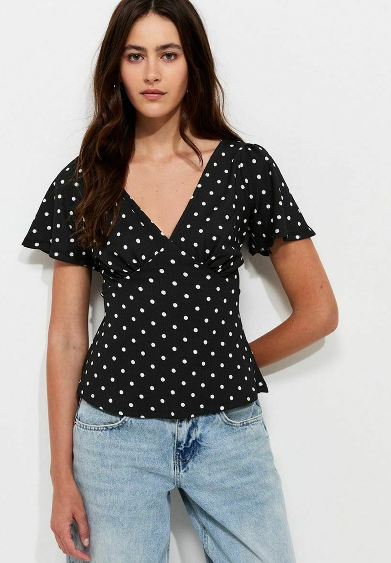 Vrouw met lang donker haar, die een zwarte blouse met V-hals en witte stippen draagt en lichtblauwe high-waisted jeans, staand tegen een witte achtergrond.