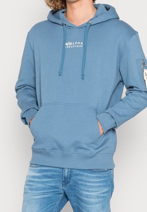Person trägt einen blauen Alpha Industries Hoodie mit Fronttasche und Reißverschlusstasche am Arm, kombiniert mit ausgewaschenen blauen Jeans. Hände in den Taschen.