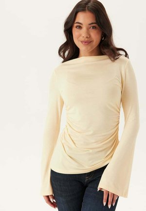 Kvinde med mørkt, bølget hår iført en cremefarvet langærmet top med rynkede sider og vidde manchetter, kombineret med mørkeblå jeans.