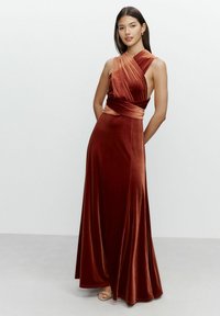 Floor-length, rust-colored velvet dress with a twisted bodice and a fitted waist. Features an asymmetrical neckline and flowing skirt.
