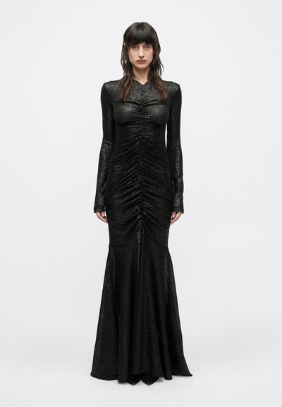 Woman standing wearing a long, fitted black sequin evening gown with ruched detailing and long sleeves against a plain gray background.