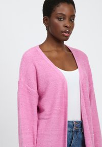 Roze cardigan met een losse pasvorm, textuur breiwerk en metallic vlokken. Heeft een diepe V-hals en wijde mouwen. Gedragen over een witte top.