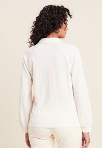Weißer strukturierter Pullover mit Kragen, ausgestattet mit Raglanärmeln und elastischen Bündchen. Die Rückansicht zeigt eine lockere Passform und glattes Material.