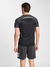 Mann, der mit dem Gesicht wegschaut und schwarze Sportshorts sowie ein kurzärmeliges Shirt mit weißen Chevron-Akzenten an Ärmeln und Shorts trägt.