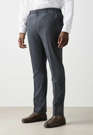 Mann trägt dunkelgraue maßgeschneiderte Hose, weißes Hemd, braune Lederloafer, steht auf hellem Boden vor weißem Hintergrund.