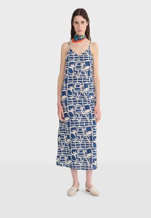 Donna che indossa un vestito midi senza maniche blu e bianco con motivi astratti, scarpe slip-on bianche e una sciarpa colorata al collo.