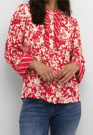 Blouse florale rouge avec des accents crème, patte de boutonnage froncée et poignets rayés, associée à un jean en denim bleu, présentée de face.