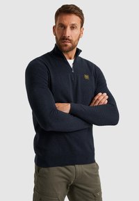 Marineblauwe sweater met ritssluiting, textuurontwerp en geribde manchetten; heeft een klein geel merklogo op de borst en een hoge kraag.