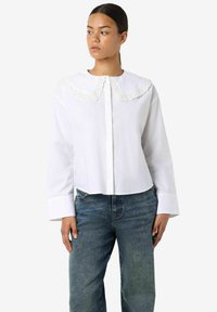 Biały krótki top z dużym, falistym koronkowym kołnierzem, długimi rękawami i zapięciem na guziki z przodu. W połączeniu z ciemnoniebieskimi jeansami.