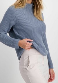 Kvinna klädd i en ljusblå stickad tröja och högmidjade beige byxor, som håller i fållen på tröjan med ena handen.