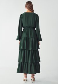 Long green dress with tiered ruffles, long sleeves, and a waist tie. Lightweight fabric and smooth texture. Worn with white heeled sandals.