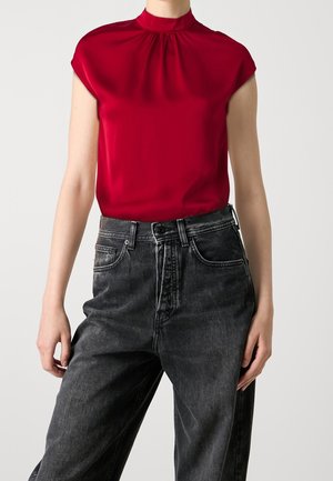 Personne portant un chemisier rouge sans manches à col montant et un jean noir délavé taille haute sur un fond blanc uni.