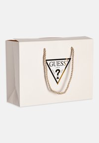 Bolsa de papel blanca con un asa de torsión dorada, que presenta un logo triangular negro con las palabras "GUESS" y "U.S.A." en dorado.