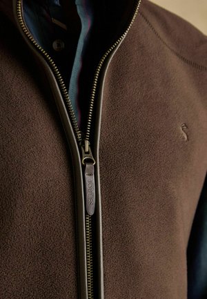 Bruine fleece jack met ritssluiting, voorzien van een soepele rits met een leren trekkoord met het label "JOULES." Inclusief een met patroon gevoerde binnenkant zichtbaar bij de kraag.