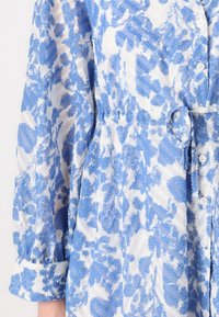 Cămașă cu un model alb și albastru, cu închidere cu butoane în față, talie strânsă și manșete răsucite. Material texturat cu un design floral.