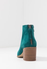 Sinihtävänvihreä mokkanahkainen nilkkuri, jossa on ribbineulekoristeinen ruskea korko ja vetoketjutakainen yksityiskohta. Sileä rakenne ja tyylikäs muotoilu.