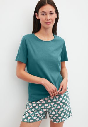 Vrouw draagt een teal shirt met korte mouwen en groene korte broek met roze madeliefjespatroon, staand tegen een effen witte achtergrond.
