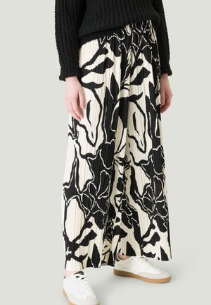 Persona con suéter negro texturizado y pantalones de pierna ancha plisados con un patrón floral abstracto en blanco y negro, combinados con zapatillas blancas.