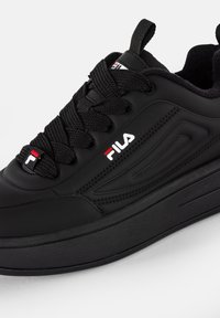 Black sneaker with textured synthetic upper, round laces, raised sole, and white Fila logo. Features a padded collar for comfort.