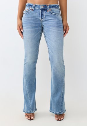 Kobieta w jasnoniebieskich jeansach bootcut i sandałach na otwartym obcasie, stoi na tle jednolitego jasnego tła, ręce luźno opuszczone wzdłuż ciała.